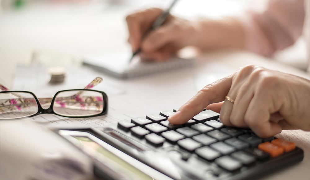 Woman using calculator and writing down number with eyeglasses in foreground